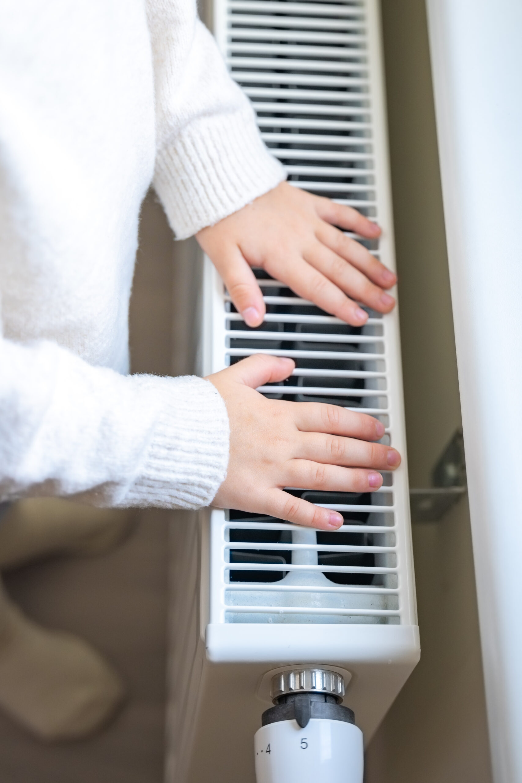 Hands of little girl in warm sweater and socks standing by window and warming up from the heating radiator. Heating in an apartment, at home.